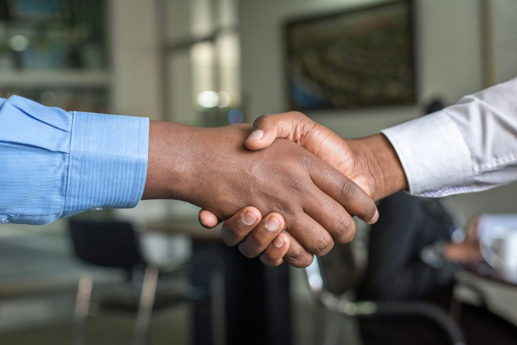 Two hands are shaking, both people are wearing long sleeve dress shirts. An office is in the background.