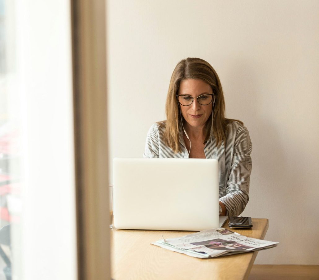 A woman with short light hair working on her laptop, she is wearing headphones. There is a phone and newspaper on a table.