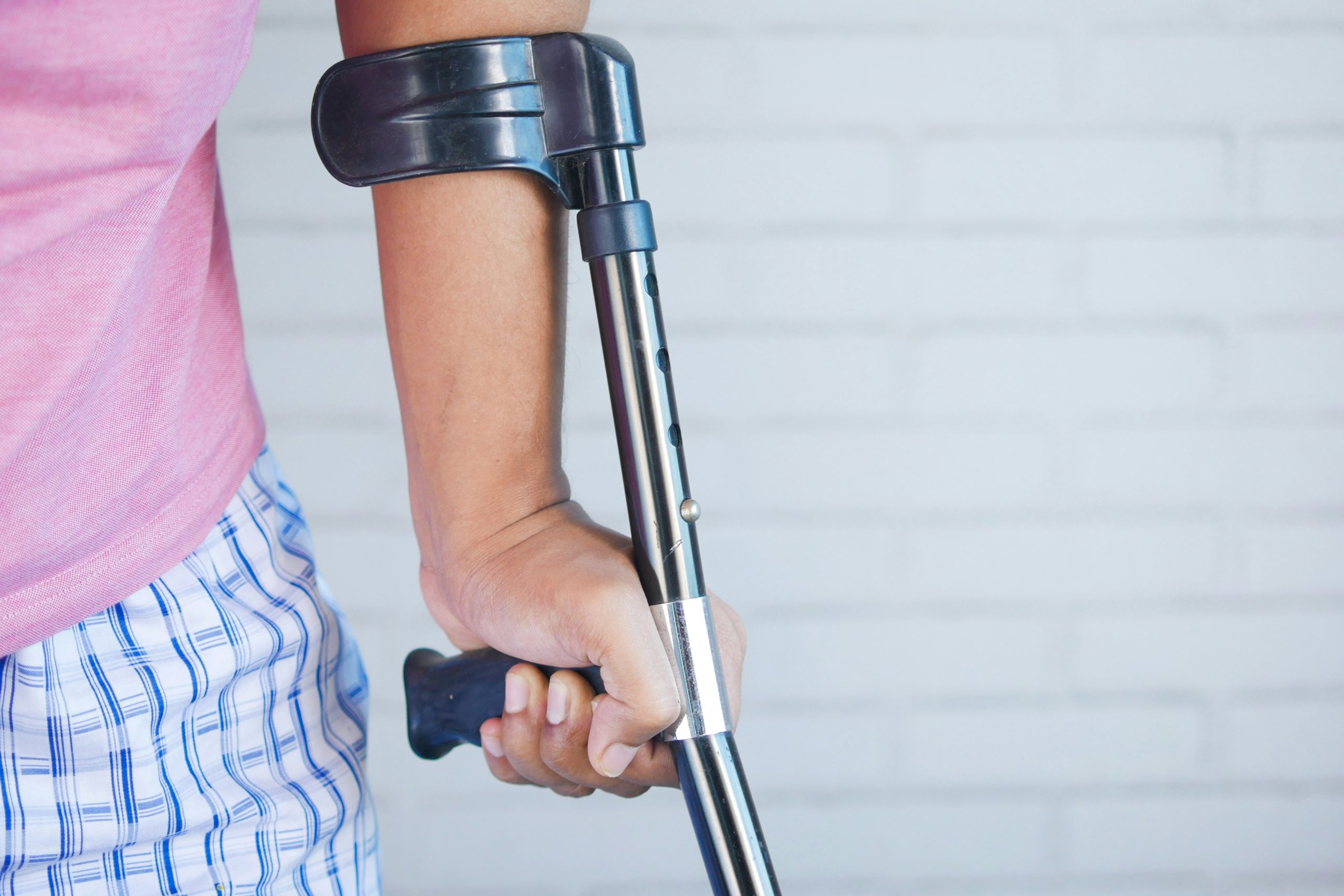 Someone using a black crutch, they have a pink top on and blue and white patterned pants. Only the side of their body can be seen.