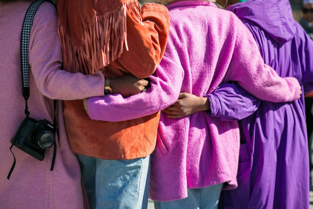 Four people with their arms linked at the waist. They are wearing colourful jackets, the photo is of their backs.