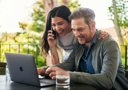 A Man and woman are sitting outside, on the table is a glass and a laptop they are both looking at. The woman has her hand around the mans shoulders and is on the fan.