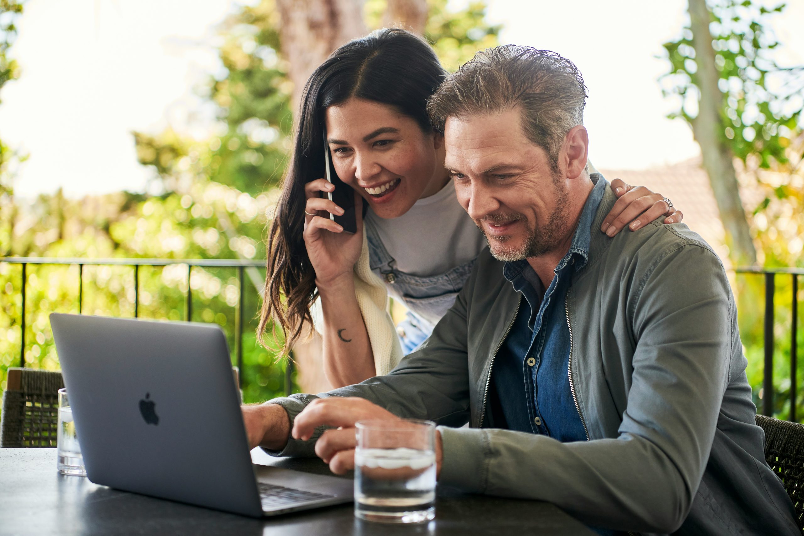 A Man and woman are sitting outside, on the table is a glass and a laptop they are both looking at. The woman has her hand around the mans shoulders and is on the fan.