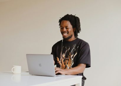 Man with black hair and beard wearing a graphic t-shirt. He is working on an apple laptop and is wearing wired headphones.