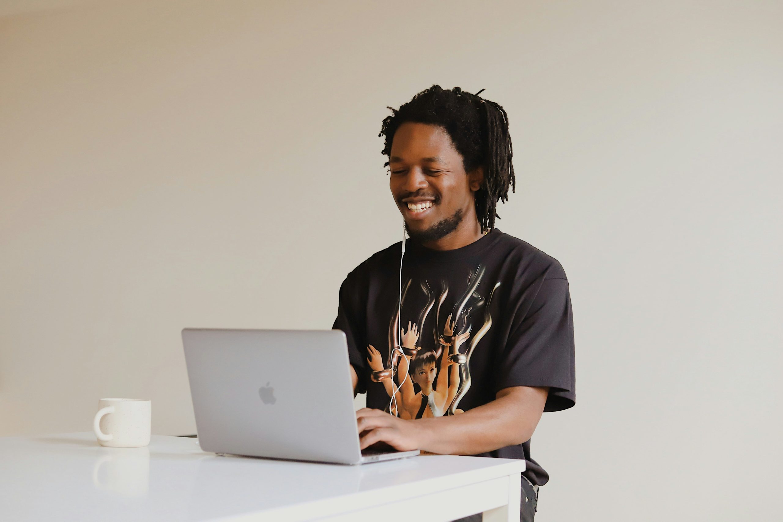 Man with black hair and beard wearing a graphic t-shirt. He is working on an apple laptop and is wearing wired headphones.