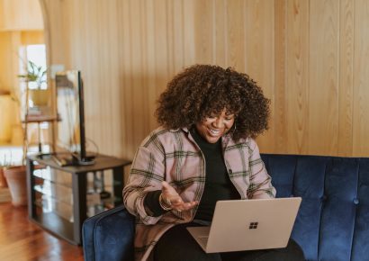 Woman with brown curly hair is smiling at her laptop which is on her lap. She is sitting on a blue couch.