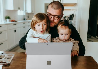 A man is sitting at a table with a computer open. There is a little girl on his lap pointing at the screen and he is holding a baby.