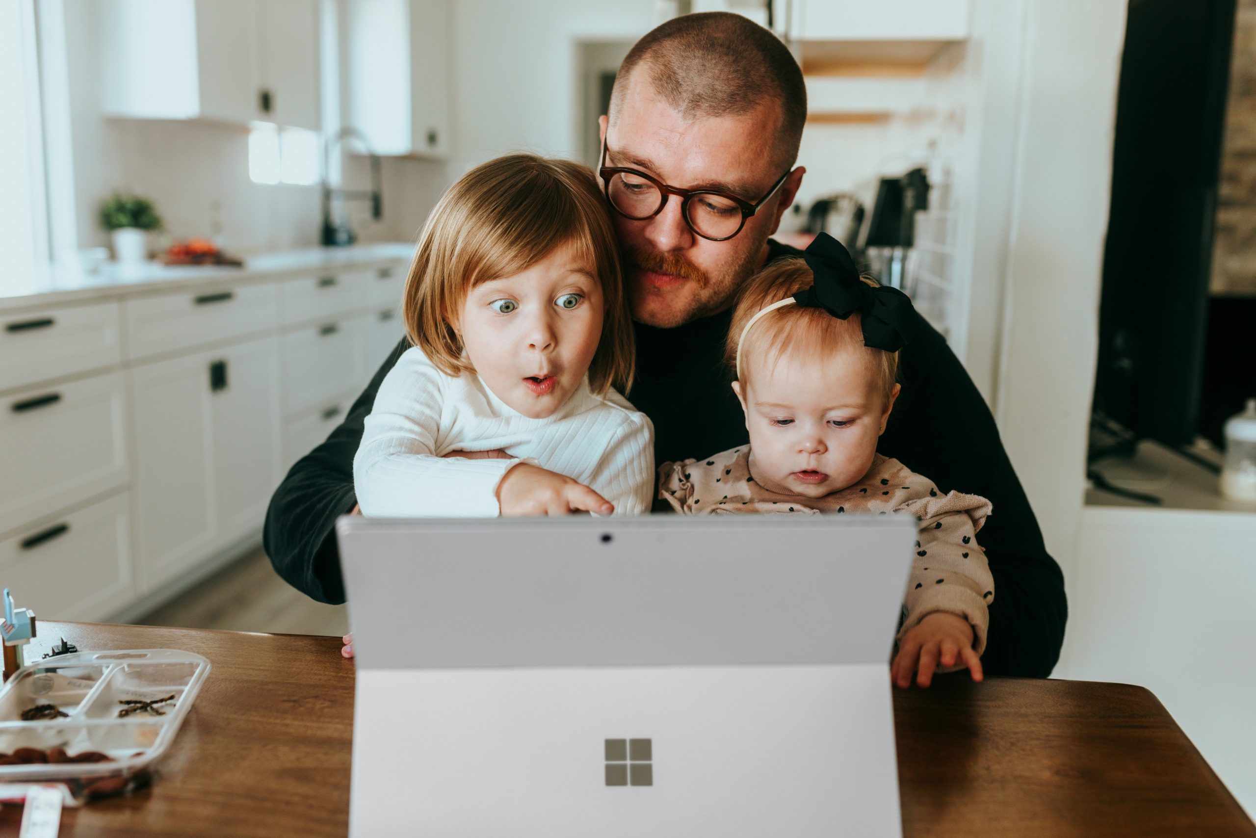 A man is sitting at a table with a computer open. There is a little girl on his lap pointing at the screen and he is holding a baby.