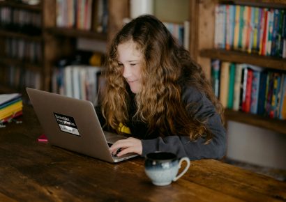 A girl with long hair is working at a table on a laptop. There is a cup on the table and there are lots of books in the background.