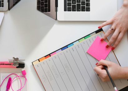 A flat lay, someone is writing on a pink post it, there is a diary, laptop and bits of stationery on the desk.