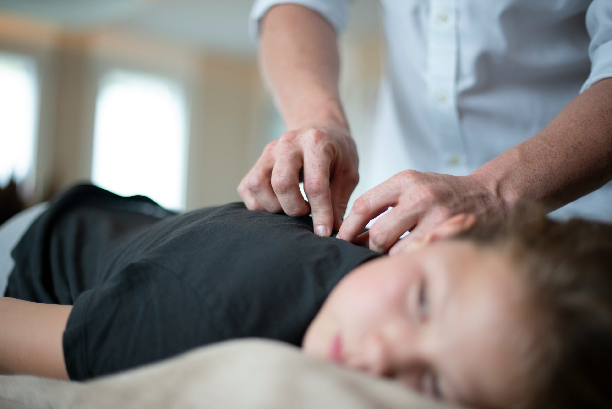 A boy in a black shirt is lying on his stomach and hands are adjusting his back.