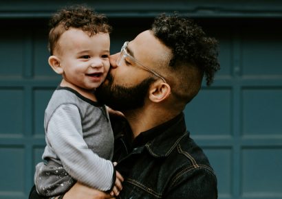 A man with curly hair, a beard and glasses is holding his baby and kissing their cheek.