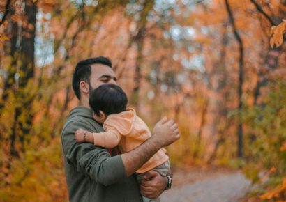 A man with dark hair is holding a baby who is wearing orange