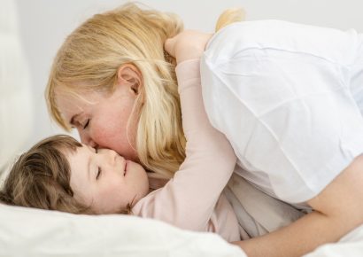A mom is playing with her daughter on the bed and kissing her cheek