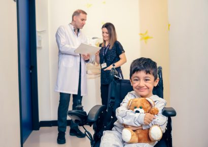 A little boy is smiling and holding a teddy he is in a wheelchair, doctors are talking behind him