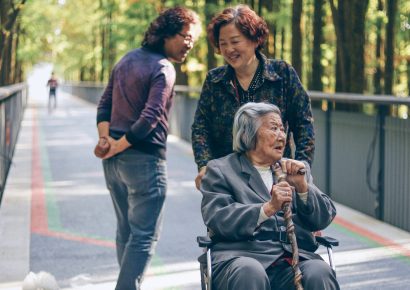 An older woman is in a wheelchair being pushed by a woman, a man is behind them with a dog