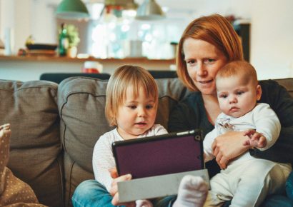 A mom sits on the couch with two young children on her lap, they are all looking at an ipad