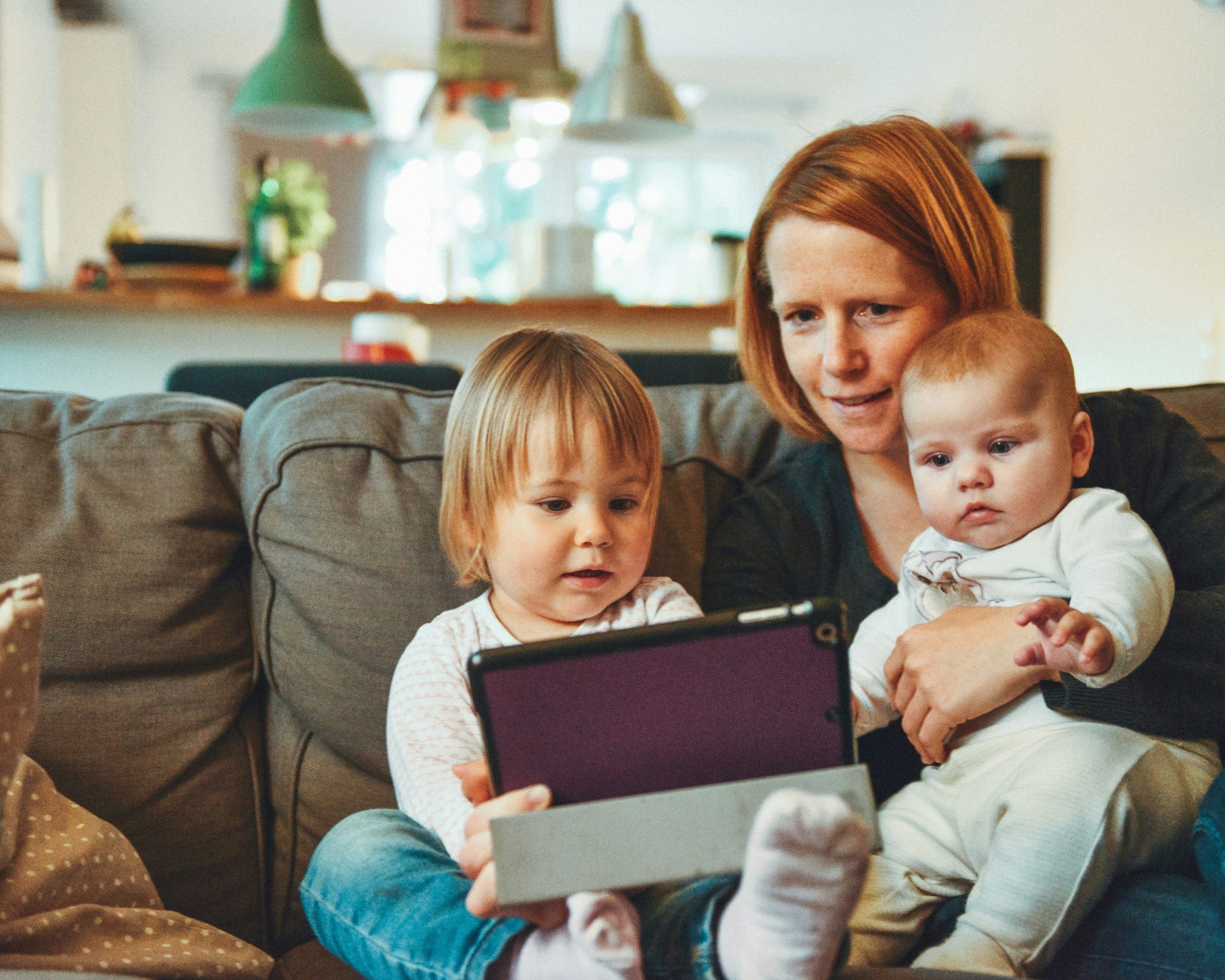 A mom sits on the couch with two young children on her lap, they are all looking at an ipad
