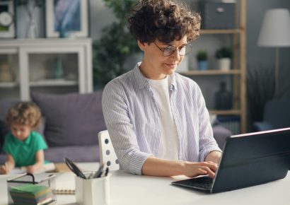 A woman is on her laptop inside a home, there are children in the back