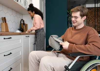 A man is drying dishes and uses a wheelchair. A woman is washing dishes in the back