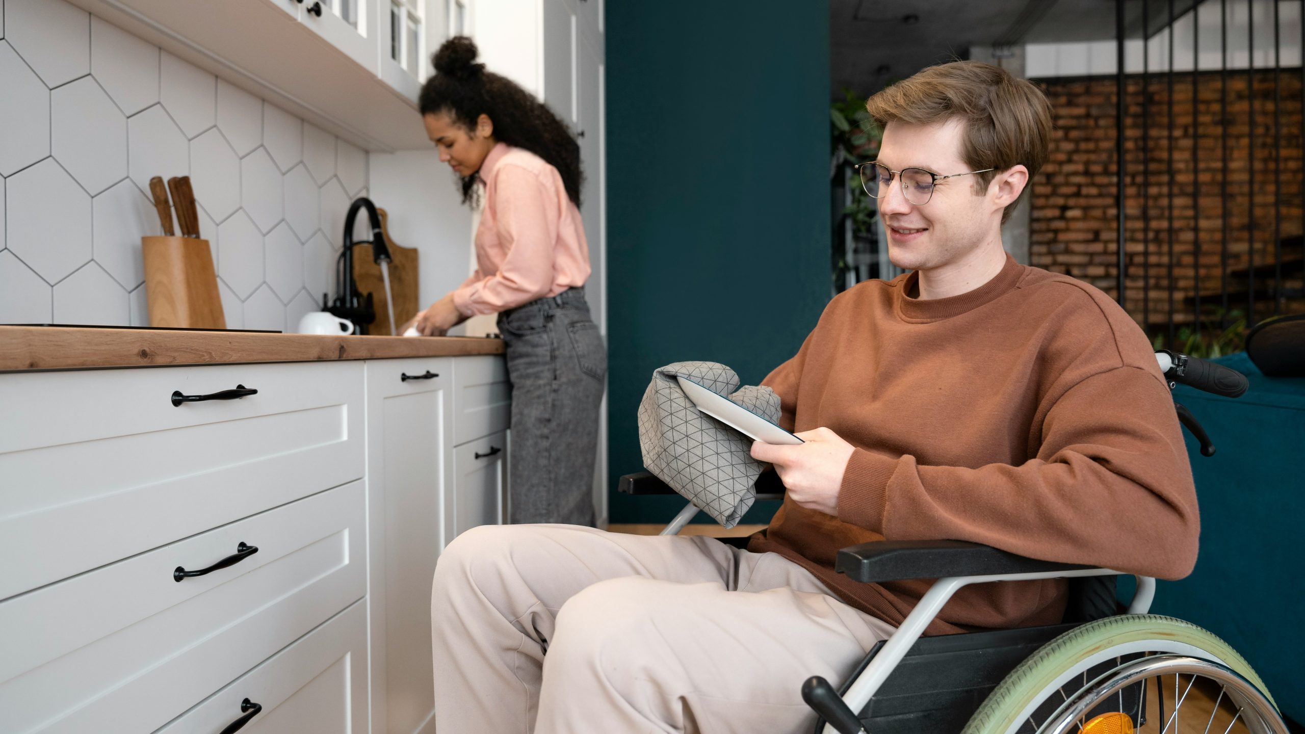 A man is drying dishes and uses a wheelchair. A woman is washing dishes in the back