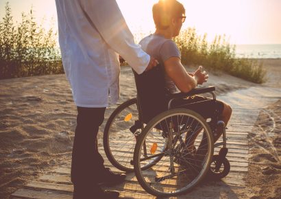 Two people are looking at the sea from the board walk at sunset. One person uses a wheelchair
