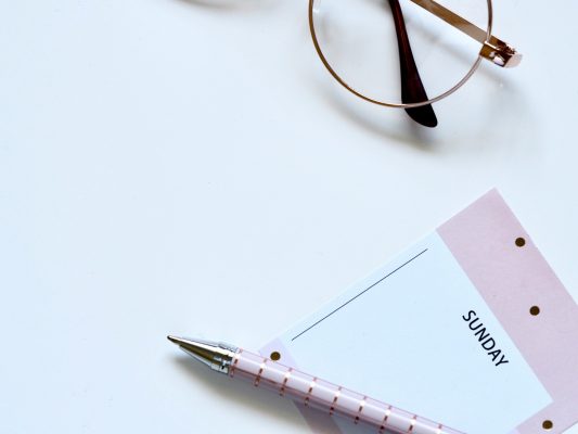 Flat lay of a notepad with 'Sunday' at the top and pen, there is a pair of thin rimmed glasses.